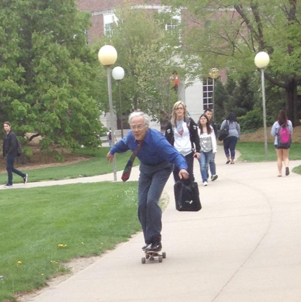 Homme âgé en skateboard roulant sur allée de campus devant groupe d'étudiants.