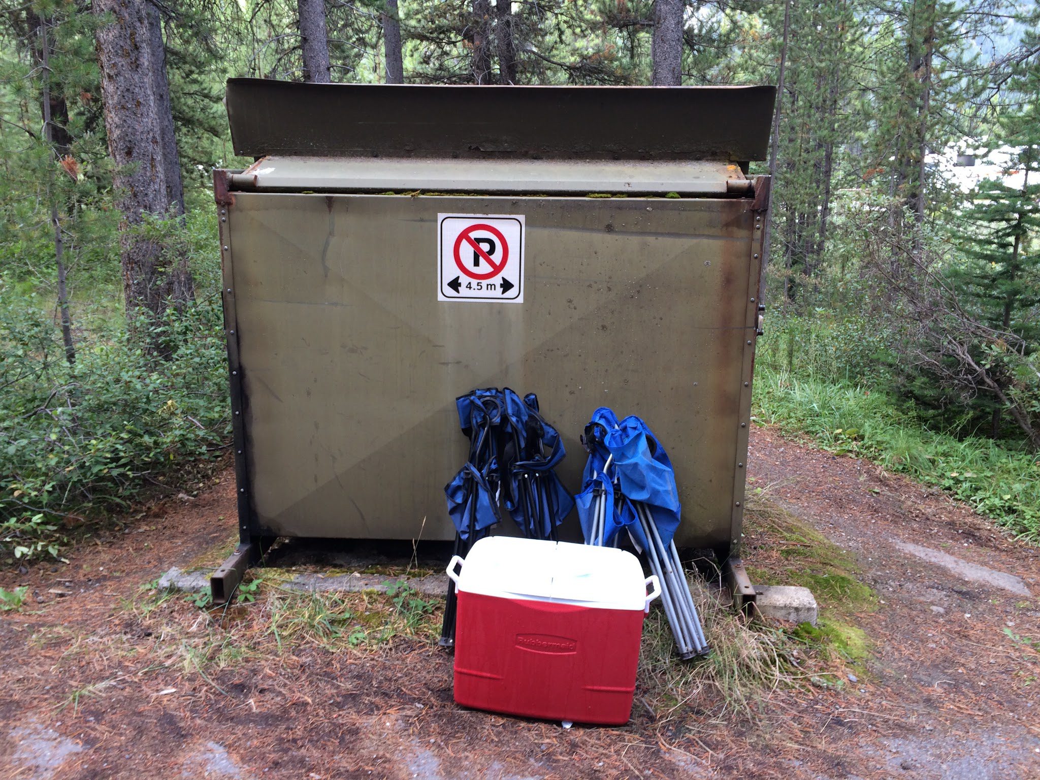 Benne de chantier grise en forêt, glacière rouge et équipement bleu, panneau interdiction