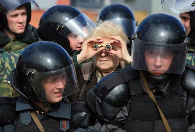 Femme âgée entourée d'officiers de police en casques noirs, geste des jumelles aux yeux