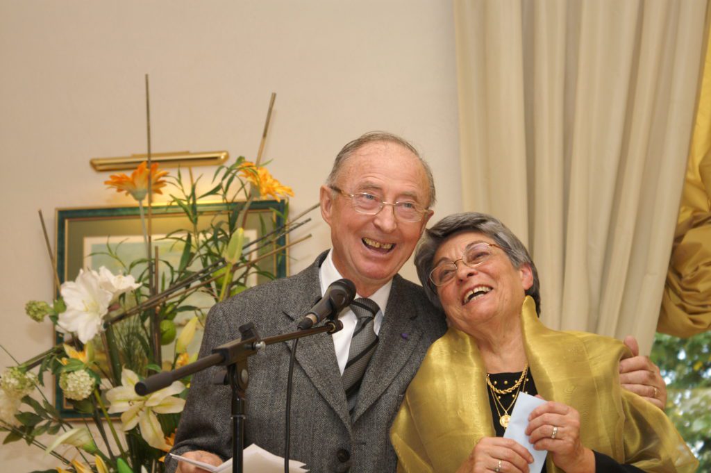 Couple âgé souriant à un micro lors d'une cérémonie, fleurs en arrière-plan