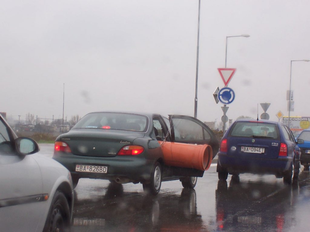 Voiture orange qui sort par la porte-coffre d'une voiture noire, dans un parking mouillé