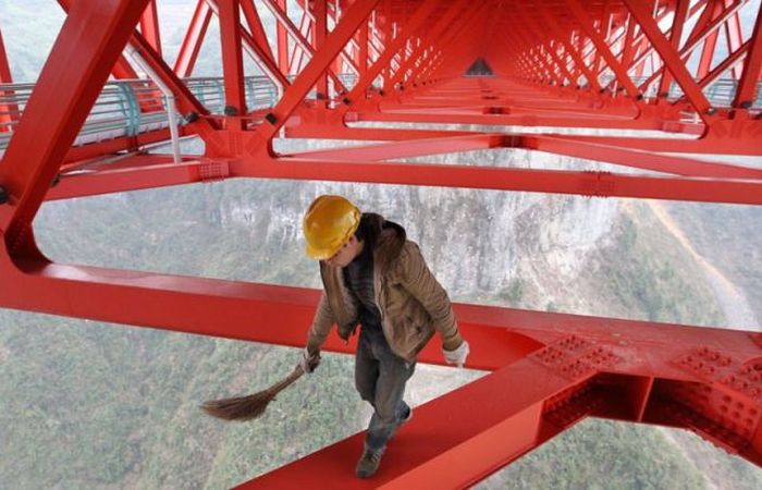 Ouvrier en casque jaune balayant structure métallique rouge géante, vue de montagne