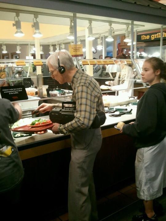 Homme âgé aux cheveux blancs portant casque audio, assis dans une cafétéria avec assiette colorée